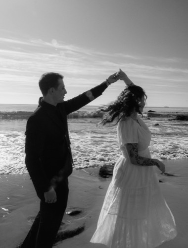 Black and white photograph of a couple dancing on the shoreline, the man turning her under his arm