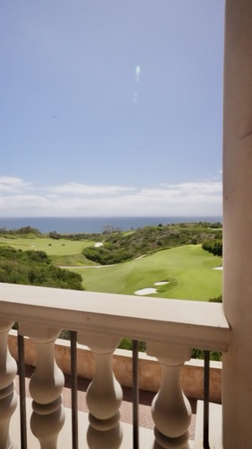 A coastal golf course and the open ocean seen from a balcony, framed by a stone balustrade